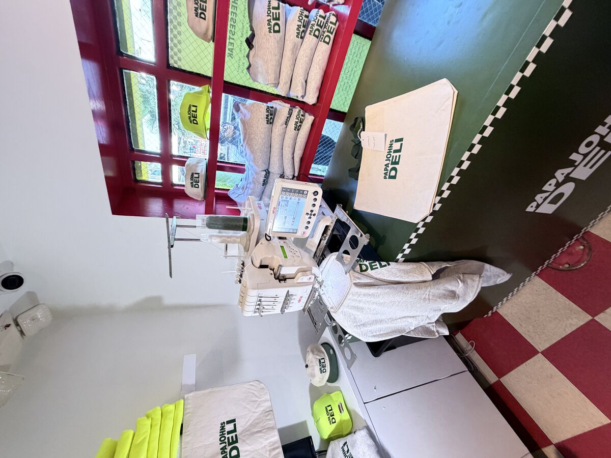 Single-head embroidery machine stitching a crewneck at a compact on-site station, with finished caps and canvas totes stacked around it