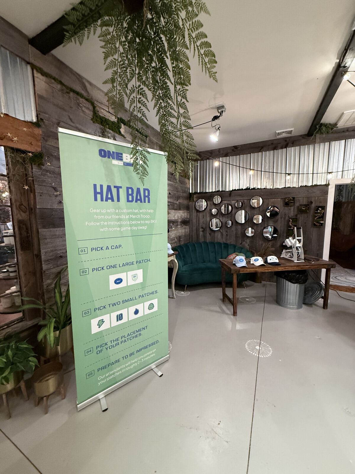 Indoor personalization lounge set up before doors open, with a step-by-step instruction banner, a press and embroidery table, and caps laid out ready