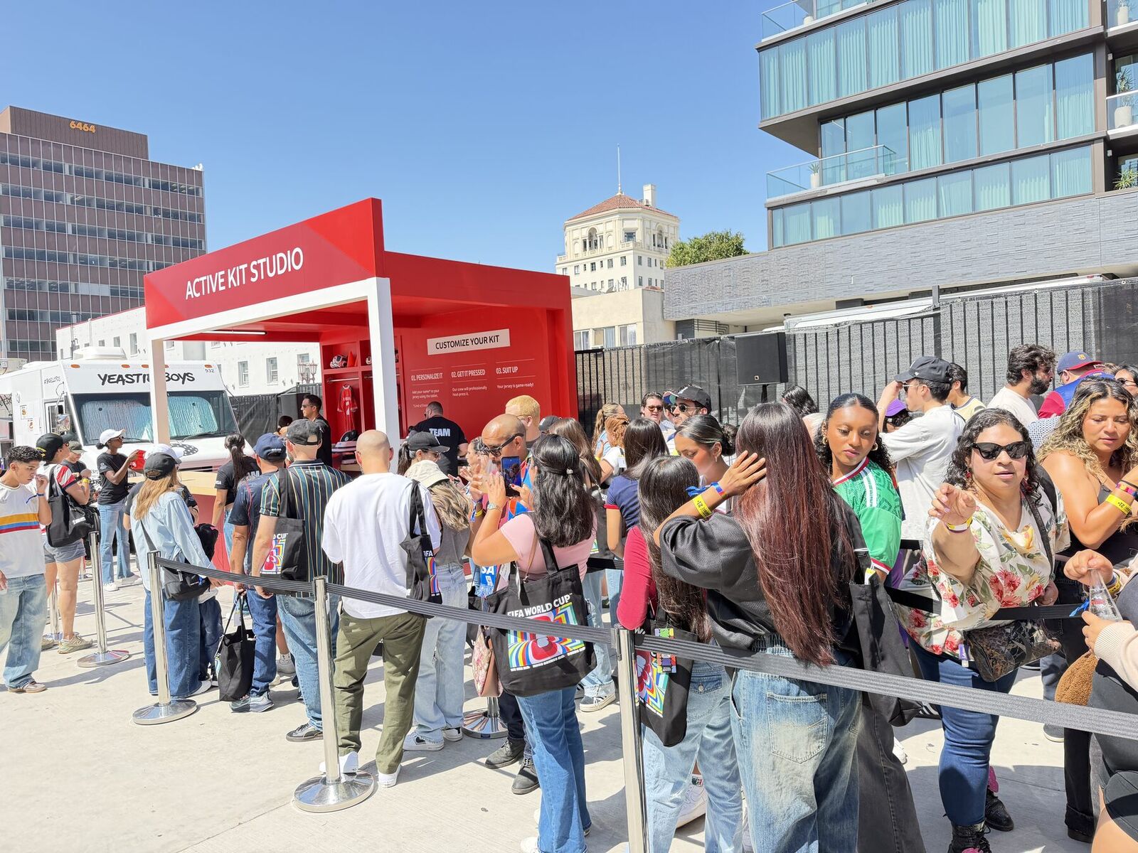 Guests waiting behind stanchions at an outdoor jersey personalization studio, with the three-step Customize Your Kit board mounted on the booth wall